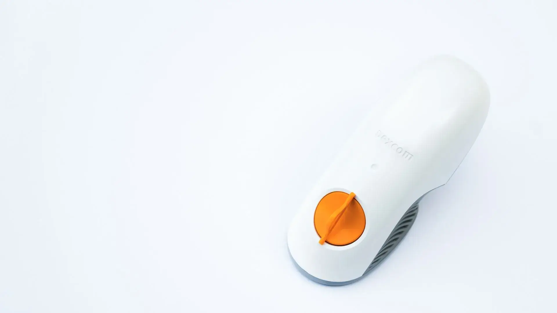 A white handheld Dexcom G6 device with a visible orange circular button or dial, placed on a plain white surface.